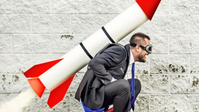 zoomed in grown man riding tricycle with large rocket on his back 